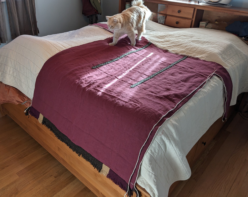 A wine colored linen tunic in the Coptic style laid out on a bed to show the shoulder stripes and fringe along the bottom. A fluffy apricot colored cat is walking on it with interest.