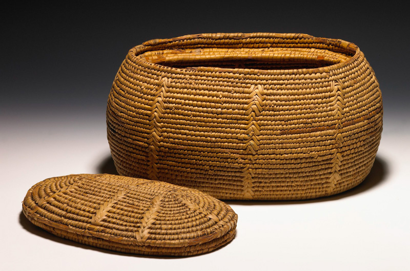 Tall elliptical basket with an inner rim made with the coiling technique. A similarly constructed lid sits next to the basket.