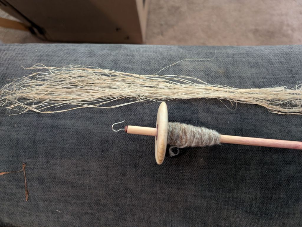 A small top-whorl spindle holding a small cone of spun wool sits on a fabric surface in front of a small hank of nettle fibers
