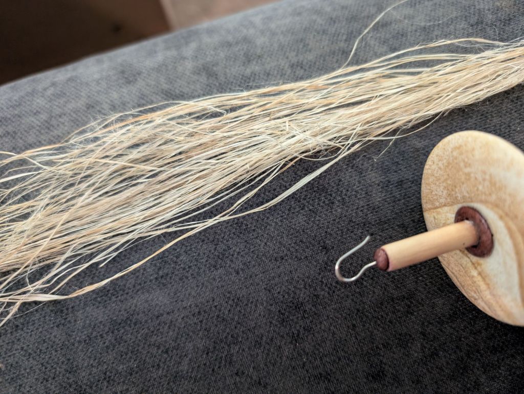 Close-up view of the prepared nettle fibers sitting on a fabric surface. The hooked top of a spindle is visible in the foreground.