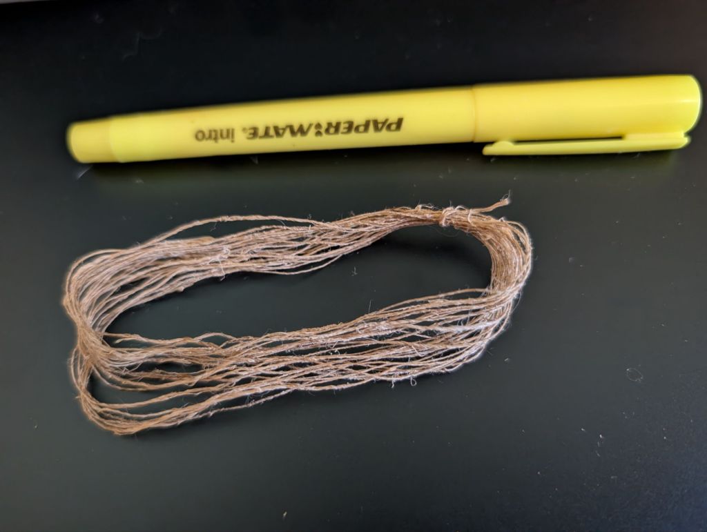 A close-up photo of a hand-sized skein of thread spun from the test hank of nettle fiber and tied in one spot is displayed in an open loop. There is a yellow highlighter pen next to it for scale; the skein is approximately three quarters the length of the highlighter.