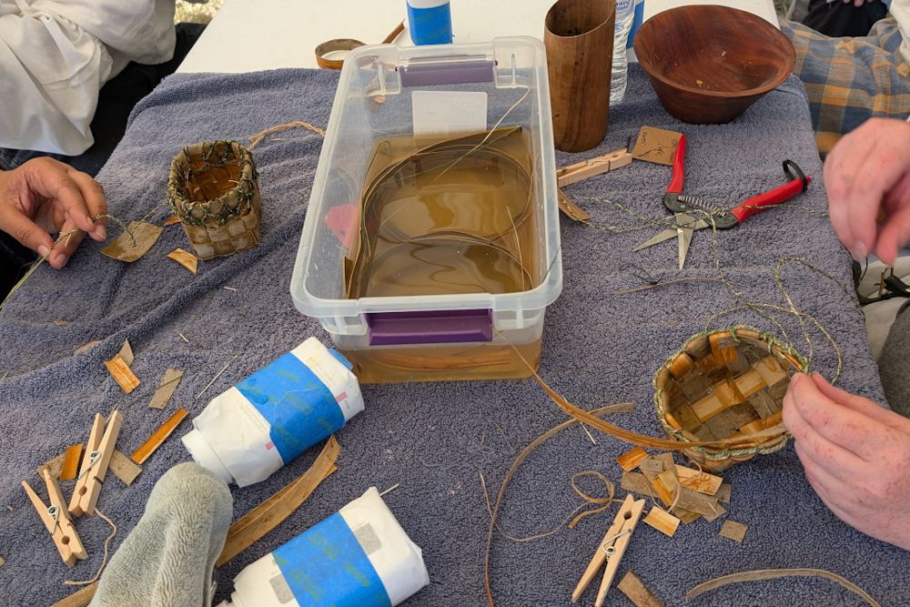 A table where students are working on making small baskets. The work surface is covered with a towel, and there is a small bin containing water and submerged pieces of willow bark in the center.