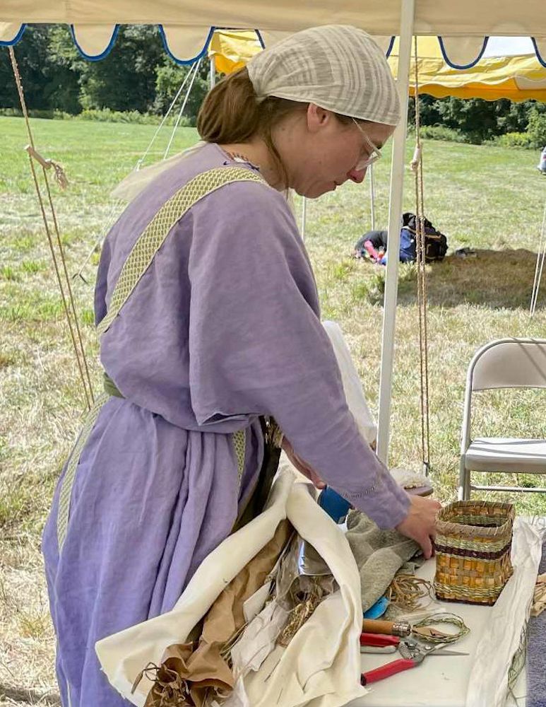 Picture of the author wearing a purple tunic and striped linen head covering. She is standing at a table that is covered with various tools, plant materials, and a completed example basket.