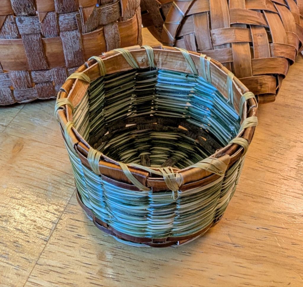 Photo of the basket with the top edge finished. A band of willow bark around the top interior edge is whip stitched into place with light green strips of black raspberry bast fiber ribbons.