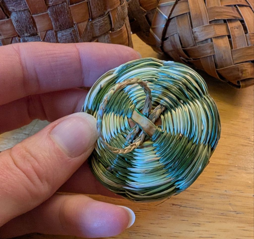 Top view of the lid, showing a thin braided loop handle attached at the center and a twined design of alternating white and green stripes radiating from the center of the lid.