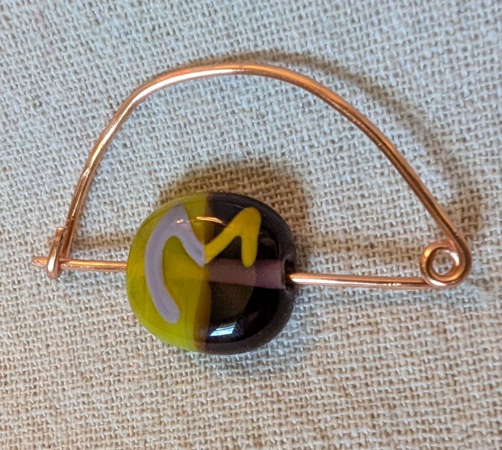 Photo of a glass bead that is a flat circle with the hole running through the circle's diameter. The circle is yellow on the left half and purple on the right half, with a symbol similar to a stylized "M" drawn on top using the opposite colors (purple on the left and yellow on the right). The bead has the pin of a simple copper bow fibula stuck through the hole
