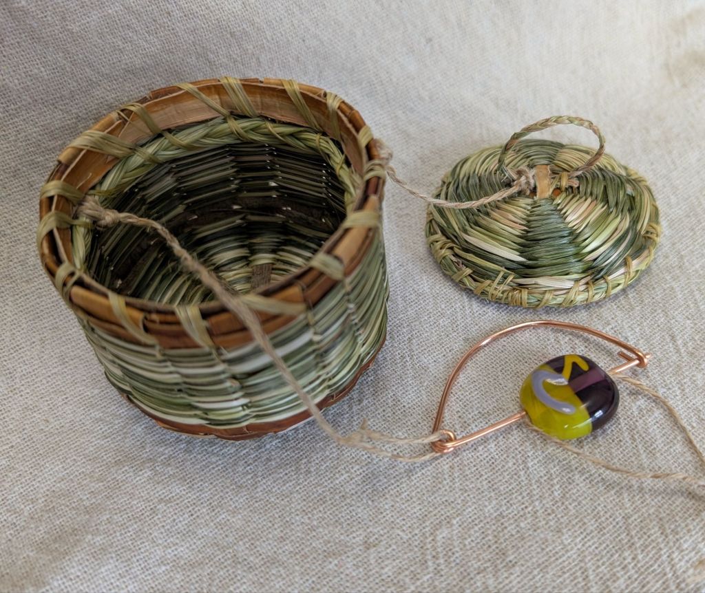 A tiny basket of red-brown willow bark and twined white and green sweetgrass, connected by cords to a matching lid and a copper pin holding a lampwork glass bead bearing the symbol of the East Kingdom's Order of the Maunche.