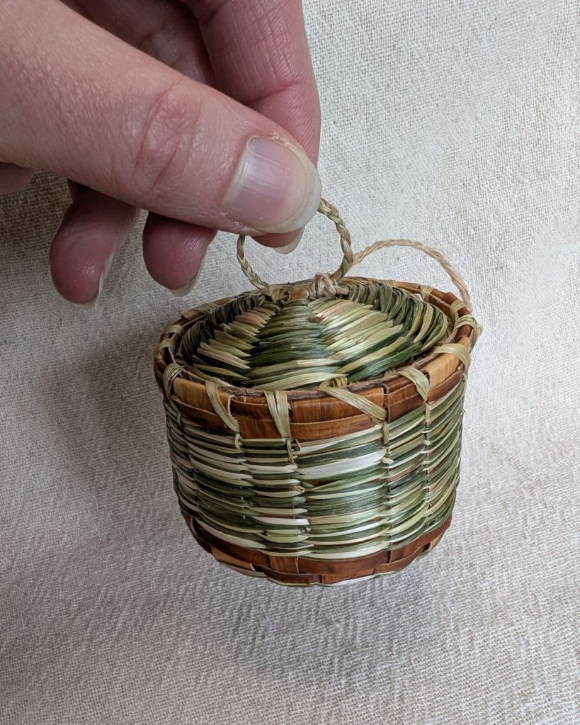 Photo of the assembled basket with the lid tucked inside, being held up in the air by only the thin braided loop on the top of the lid. A thin, off-white cord connects the side of the basket to the lid near the looped braid handle.